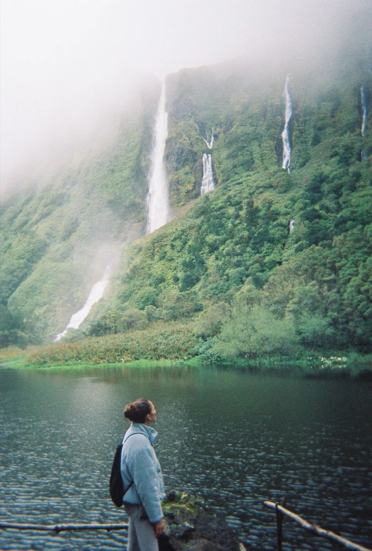 Standing at the edge of a lake in Flores, in the Azores, looking up at waterfalls falling down a green cliff.
